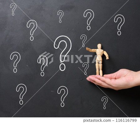 A woman's hand holding a wooden miniature puppet against a black chalkboard with chalk-drawn question marks. 116116799