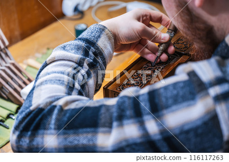 man artisan carpenter carves pattern on wooden casket with a chisel in workshop 116117263