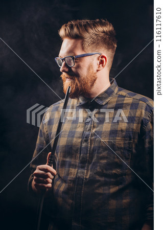 Stylish beard man in glasses and checked shirt smoking hookah at night bar. 116117610