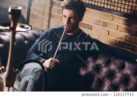 Young handsome serious bearded man smoking hookah indoors 116117713