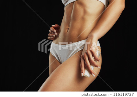 Cropped image of tanned and muscular female body with abs, in underwear against black studio background. Body cream 116119727