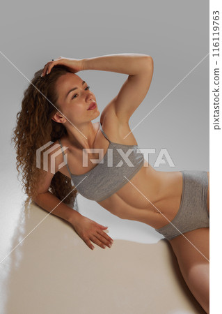 glow of health and well-being. Portrait of young beautiful woman with curly hair and slim fit body, posing in underwear against grey studio background 116119763
