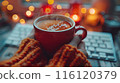 Hands Holding a Red Mug of Hot Drink With Steam in Front of a Computer Keyboard at Night 116120379