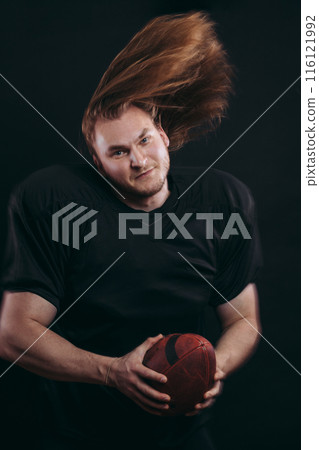 Confident american football player in black sportwear holding helmet and ball 116121992