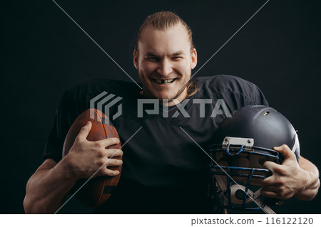 Confident american football player in black sportwear holding helmet and ball 116122120