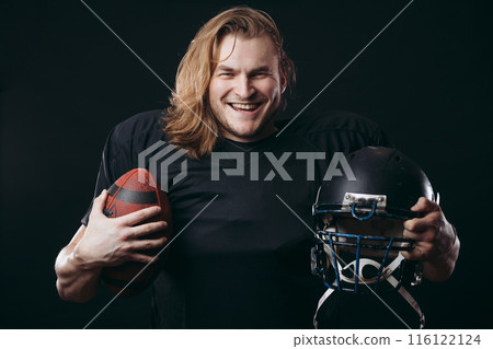 Confident american football player in black sportwear holding helmet and ball 116122124