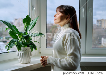 Middle aged woman with closed eyes enjoying winter sun at home near window Middle aged woman with closed eyes enjoying winter sun at home near window 116122292