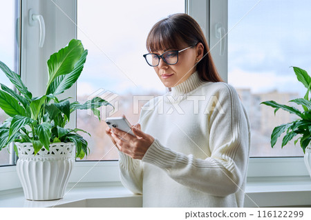 Calm confident middle aged woman using smartphone, at home near window Calm confident middle aged woman using smartphone, at home near window 116122299