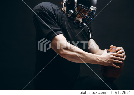 American football player with helmet and armour with a ball against black wall American football player with helmet and armour with a ball against black wall 116122695