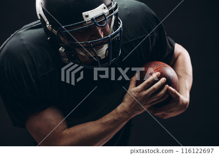 American football player with helmet and armour with a ball against black wall American football player with helmet and armour with a ball against black wall 116122697