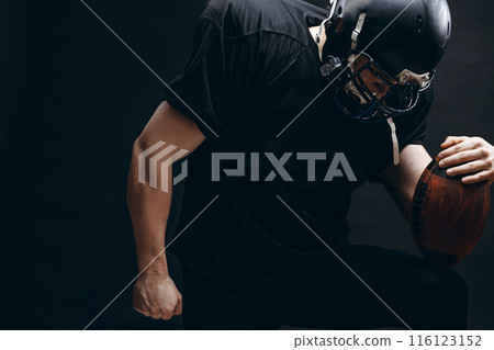 American football player with helmet and armour with a ball against black wall American football player with helmet and armour with a ball against black wall 116123152