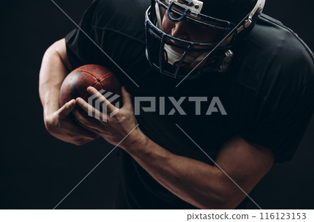 American football player with helmet and armour with a ball against black wall 116123153