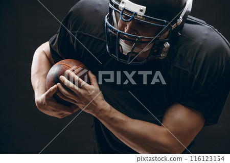 American football player with helmet and armour with a ball against black wall American football player with helmet and armour with a ball against black wall 116123154