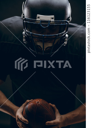American football player with helmet and armour with a ball against black wall American football player with helmet and armour with a ball against black wall 116123155