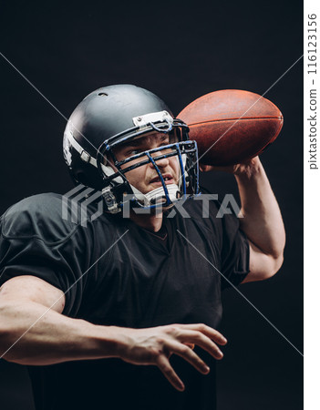American football player with helmet and armour with a ball against black wall American football player with helmet and armour with a ball against black wall 116123156
