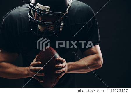 American football player with helmet and armour with a ball against black wall American football player with helmet and armour with a ball against black wall 116123157