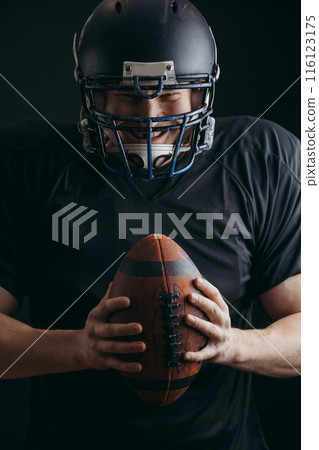 American football player in black sportwear with a ball on black background 116123175