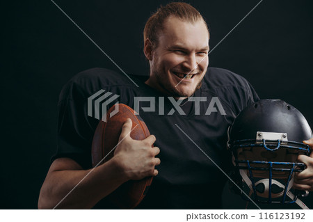 Confident american football player in black sportwear holding helmet and ball 116123192