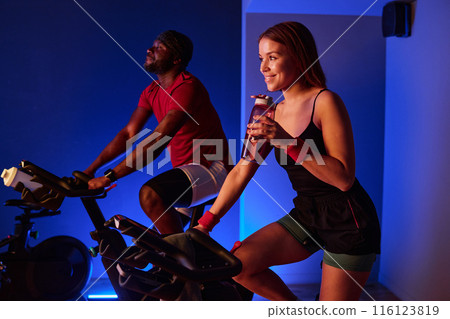 Biracial couple doing cardio in cardio neon zone of premium fitness club, woman smiling widely while she drinking BCAA, and man smiling while riding exercise bike 116123819