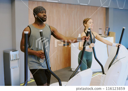 Biracial gym residents running on elliptical machine, pretty woman enjoying process, and young African American man listening to music 116123859