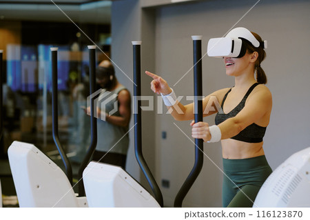 Young woman playing VR game and smiling widely while she doing cardio, African American man surfing Internet in background Young woman playing VR game and smiling widely while she doing cardio, African American man surfing Internet in background 116123870