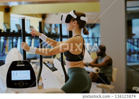Young woman doing cardio on elliptical machine and playing VR game, while African American man doing leg superset on leg extension in background 116123874