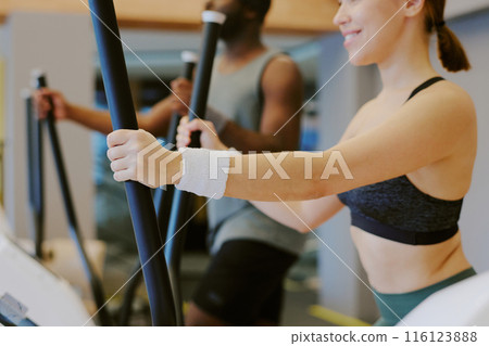 Medium close up of strong and toned female arms holding elliptical machine handles, African American man doing cardio as well in background 116123888