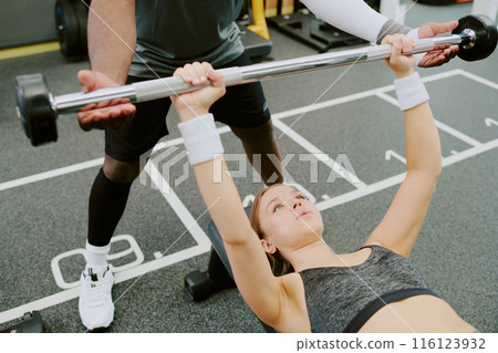 Female client doing bench press while her African American male coach spotting her Female client doing bench press while her African American male coach spotting her 116123932