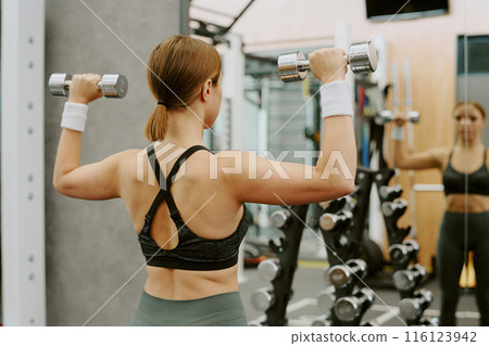 Woman looking in mirror and watching her exercise technique while she doing dumbbell shoulder press 116123942