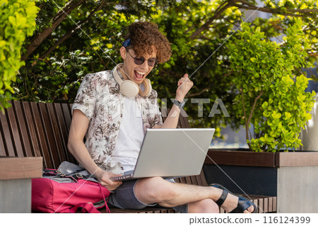 Happy man tourist using laptop computer celebrating win good message news sitting on bench in city 116124399