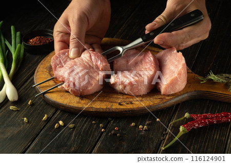 The cook hands preparing a meat steak for grilling. Chef hands skewering raw veal meat with a fork on the kitchen counter. Advertisement space on black table 116124901