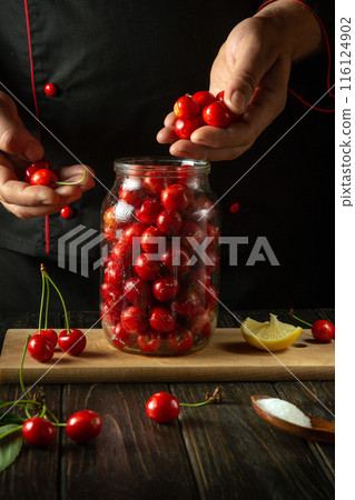 The cook fills the jar with cherries. The process of making a sweet drink from fresh berries and sugar on the kitchen table. Preserving cherries in a jar The cook fills the jar with cherries. The process of making a sweet drink from fresh berries and sugar on the kitchen table. Preserving cherries in a jar 116124902