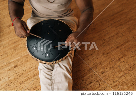 Top view of unrecognizable African male in trance playing meditative instrument tank drum using sticks sitting in meditation room. Concept of mindfulness, tranquility, harmony, sound therapy. 116125246