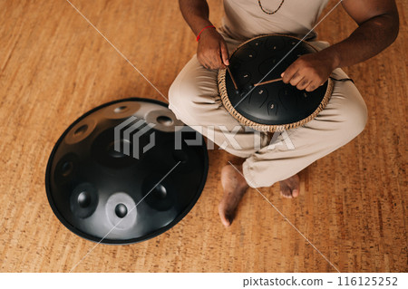 High-angle view of unrecognizable black male in trance playing meditative instrument tank drum using sticks sitting in meditation room. Concept of mindfulness, tranquility, harmony, sound therapy. 116125252