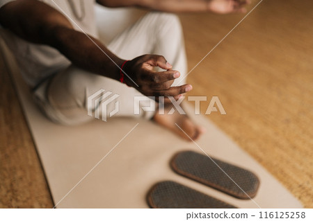 Closeup cropped shot of unrecognizable black male meditating in lotus pose at home sitting on yoga mat by sadhu board. African man prepares for practice. Concept of mindfulness, tranquility, harmony. 116125258