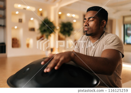 Closeup of African drummer in trance drumming rhythm on steel tongue drum by hands sitting on floor in lotus pose with closed eyes. Relaxed black male playing ethnic music using glucophone. 116125259