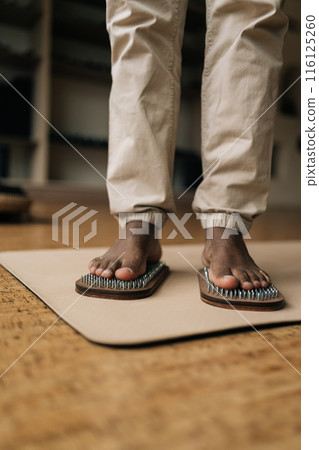 Vertical cropped shot of unrecognizable African man standing on wooden yoga Sadhu Board with metal nails for practice and meditation at home. Concept of alternative medicine and recreational activity. 116125260
