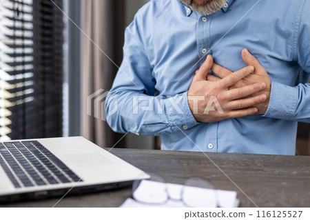 A middle-aged man in a blue shirt clutches his chest in pain while working on a laptop in a professional office setting. The image conveys stress, discomfort, and potential heart health issues. A middle-aged man in a blue shirt clutches his chest in pain while working on a laptop in a professional office setting. The image conveys stress, discomfort, and potential heart health issues. 116125527