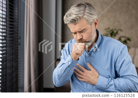 Elderly man with grey hair and beard coughing and holding chest with painful expression. Health concept of illness, bronchitis, lung disease, or flu. Indoors with window and blurred background. 116125528