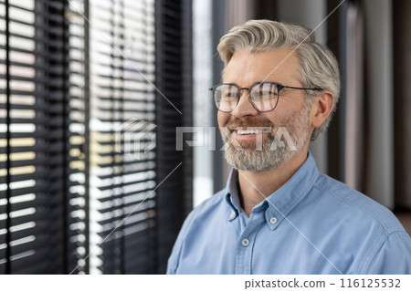 Mature man with glasses smiling while looking out window with blinds in contemporary office environment. Natural light illuminating cheerful expression capturing professional yet relaxed demeanor Mature man with glasses smiling while looking out window with blinds in contemporary office environment. Natural light illuminating cheerful expression capturing professional yet relaxed demeanor 116125532