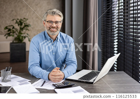 Middle-aged businessman in blue shirt smiling at his desk while working on a laptop. The modern office setup includes documents, a calculator, and natural light Middle-aged businessman in blue shirt smiling at his desk while working on a laptop. The modern office setup includes documents, a calculator, and natural light 116125533
