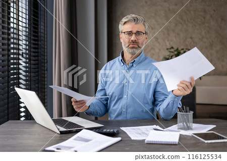 Professional businessman sitting at desk holding documents, displaying concern while working on laptop in modern office. Concept of work stress, business finance, office environment 116125534
