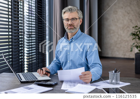 Middle-aged businessman wearing glasses and a blue shirt, analyzing documents at his desk in a modern office. He appears confident and professional, with a laptop, calculator, and paperwork Middle-aged businessman wearing glasses and a blue shirt, analyzing documents at his desk in a modern office. He appears confident and professional, with a laptop, calculator, and paperwork 116125535