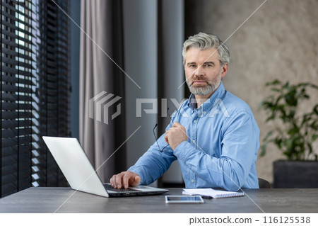 Confident mature man working from home office using laptop. Professional setting with natural light through window blinds. Modern workplace environment reflecting focus and productivity. 116125538