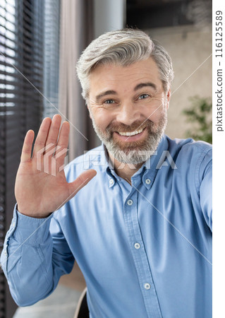A cheerful mature office worker waves at the camera, greeting on a video call. Wearing a blue shirt, he presents a warm and friendly demeanor in a professional office setting A cheerful mature office worker waves at the camera, greeting on a video call. Wearing a blue shirt, he presents a warm and friendly demeanor in a professional office setting 116125589