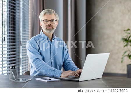 Confident professional man with glasses working on laptop in modern office setting. Workspace includes notebook, pen, tablet. Portrays productivity, focus, technology, and business environment. Confident professional man with glasses working on laptop in modern office setting. Workspace includes notebook, pen, tablet. Portrays productivity, focus, technology, and business environment. 116125651