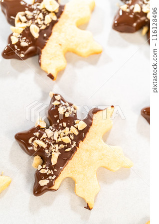 Creating cutout sugar cookies, partially dipped in chocolate and topped with hazelnut pieces, placed on parchment paper. Creating cutout sugar cookies, partially dipped in chocolate and topped with hazelnut pieces, placed on parchment paper. 116128496