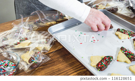 Carefully packaging Christmas cutout cookies, half-dipped in chocolate and presented in clear cellophane wrapping, perfect for festive gifting. 116128651