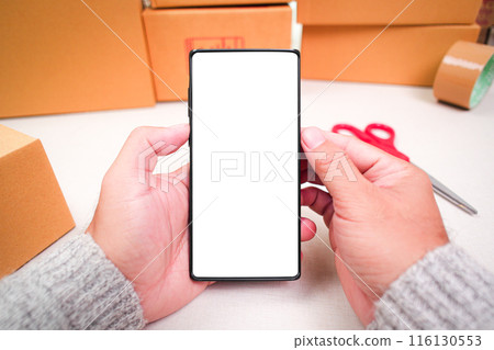 Mobile phone with blank white screen in a human hand and cardboard boxes on desk for product packing , copy space for text Mobile phone with blank white screen in a human hand and cardboard boxes on desk for product packing , copy space for text 116130553