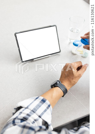 A senior biracial male is viewing a tablet with a blank screen at home, holding medicine, copy space 116130621